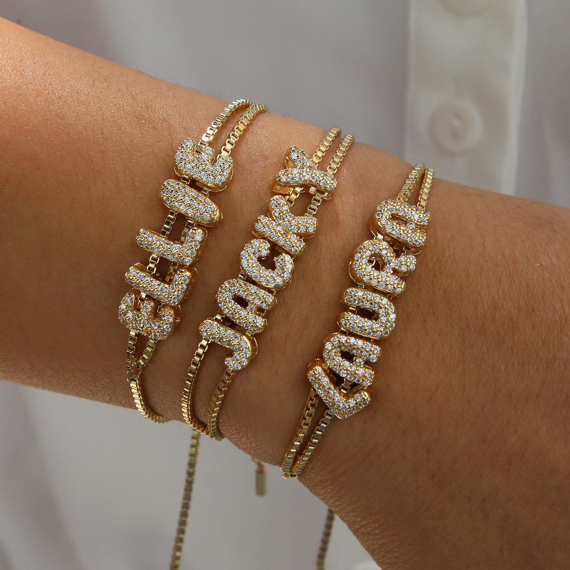 Gold name bracelets with diamonds on a wrist against a neutral background